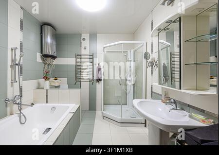 Ein gut beleuchtetes Badezimmer verfügt über eine Badewanne, eine Glasduschkabine, ein Standwaschbecken mit einem Spiegelschrank und einen Wasserheizer, mit einem gefliesten Boden und Wänden Stockfoto