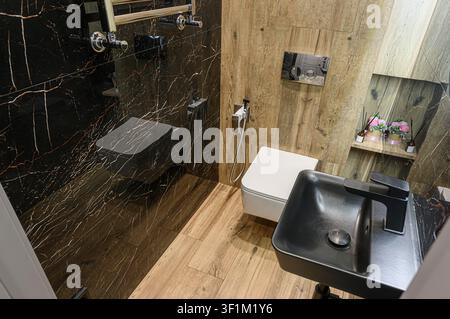 Stilvolles Badezimmer mit schwarzen Marmorwänden, hellen HolzAkzenten, modernen Armaturen einschließlich eines schwarzen Waschtischs, einer Toilette und einem beheizten Handtuchhalter Stockfoto