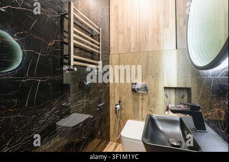 Stilvolles Badezimmer mit schwarzen Marmorwänden, hellen HolzAkzenten, modernen Armaturen einschließlich eines schwarzen Waschtischs, einer Toilette und einem beheizten Handtuchhalter Stockfoto