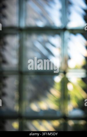 Vertikale unscharfe abstrakte Aufnahme eines Glasblockfensters, mit weichem Sonnenlicht, diagonalen Schatten und warmen Bokeh-Helligkeitsflecken mit blauen Nuancen Stockfoto