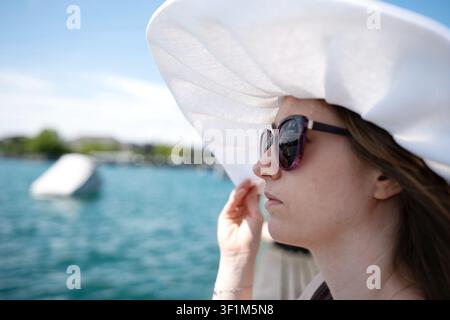 Eine Nahaufnahme im Profil einer jungen Frau, die einen großen weißen Sonnenhut und eine violette Sonnenbrille trägt und den Rand ihres Hutes hält, aufgenommen im Freien von b Stockfoto