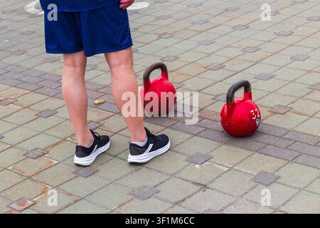 Niedrige Ansicht eines Athleten, der während eines Outdoor-Fitnesswettbewerbs auf einem gepflasterten Platz neben zwei schweren roten 32-kg-Kettlebells steht. Stockfoto