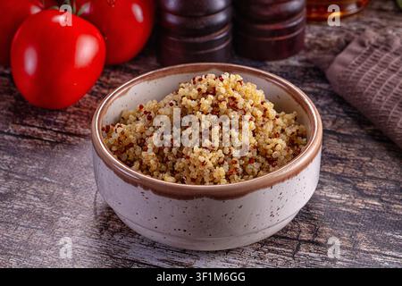 Nahaufnahme von gekochter Quinoa in einer gesprenkelten Keramikschale mit roten Tomaten, hölzernen Salz- und Pfefferstreuern auf einer verwitterten Holzoberfläche. Warme natürliche Beleuchtung hebt Stockfoto