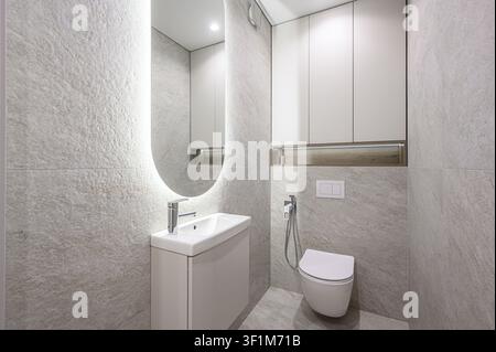 modernes Badezimmer mit strukturierten grauen Fliesen, einer weißen Waschtischschrank mit Waschbecken, einem beleuchteten ovalen Spiegel und einer wandmontierten Toilette. Stauraumschränke sind Stockfoto