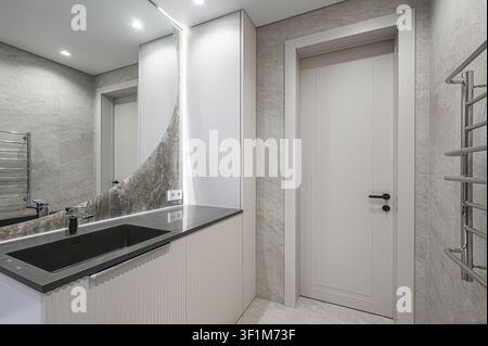 modernes Badezimmer mit einem schwarzen Waschtisch, einem gebogenen Spiegel, einer weißen Tür und grauen Fliesenwänden Stockfoto