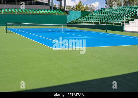 Leerer Außen-Tennisplatz mit Reihen von grünen Zuschauersitzen unter einem strahlend blauen Himmel. Sportstätte mit offenen Tribünen und farbenfroher Spielfläche Stockfoto
