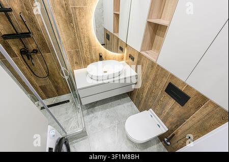 Stilvolles Badezimmer mit Fliesen in Holzoptik, einem modernen Waschbecken mit beleuchtetem Rundspiegel und einer Glasdusche. Graue Fliesenböden und weiße Armaturen runden das Ganze ab Stockfoto