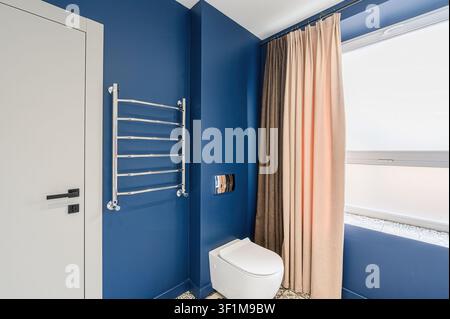 Modernes Badezimmer mit blauen Wänden, einer weißen Toilette, Handtuchhalter, weißer Tür und Vorhängen Stockfoto