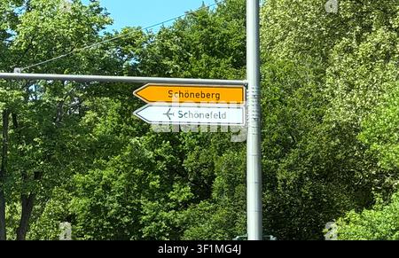 Richtungsschild, das Schoneberg und Schonefeld anzeigt, mit grünen Bäumen im Hintergrund an einem sonnigen Tag in einem städtischen Gebiet Stockfoto