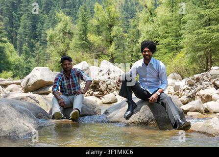 Zwei junge indische männliche Freunde sitzen zusammen auf Felsen in einem klaren Bergbach, lächeln und schauen in die Kamera, während sie einen entspannten Moment genießen. Stockfoto