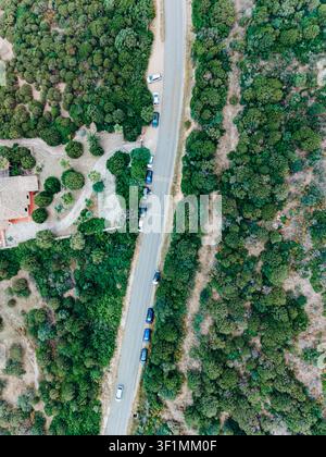 Luftaufnahme einer schmalen Straße, die einen dichten Wald durchquert, mit mehreren geparkten Autos und einem Wohnhaus, das von Bäumen umgeben ist. Das Bild stellt tra Stockfoto
