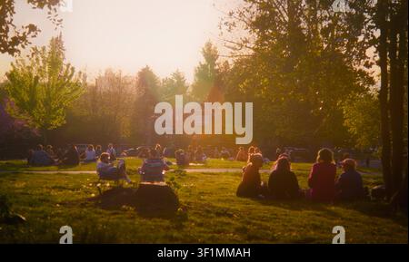 Menschen entspannen sich und sitzen auf dem Gras während eines dunstigen goldenen Sonnenuntergangs in Grandview Park auf Commercial Drive in Vancouver, British Columbia, Kanada. Stockfoto