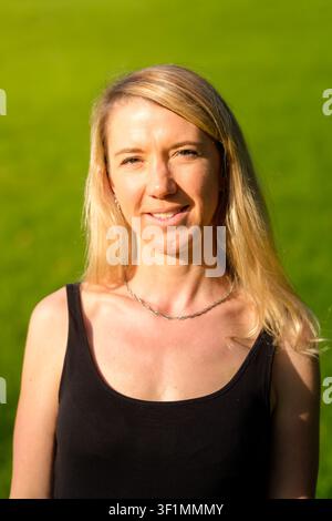 Blonde Frau lächelt, steht im Freien auf einer grünen Wiese bei natürlichem Licht Stockfoto