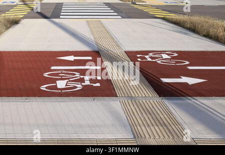 Fahrradsymbol-Straßenmarkierung auf einem roten Fahrstreifen entlang eines Betonrandsteins mit einer rutschfesten Gehwegabdeckung. Zebrastreifen, Straße und Parkplatz davor. Stockfoto