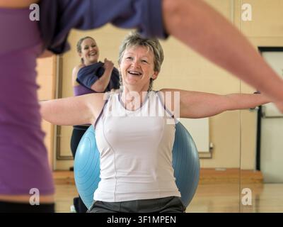 Reife Frau trainiert im Fitnessstudio mit Fitnessball, während eine junge Frau sich auf das Training vorbereitet Stockfoto