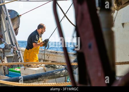Kommerzieller Fischer in gelben wasserdichten Latzhosen zieht ein Seil auf dem Arbeitsdeck eines Fischerboots, mit sichtbar werdenden Takelungen und Ausrüstungen vor dem blauen Ope Stockfoto