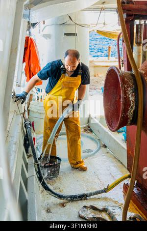 Kommerzieller Fischer in gelben wasserdichten Overalls, der das Deck eines arbeitenden Fischerboots mit einem Schlauch auf See wäscht, mit sichtbarer Ausrüstung, Seil und Winde Stockfoto