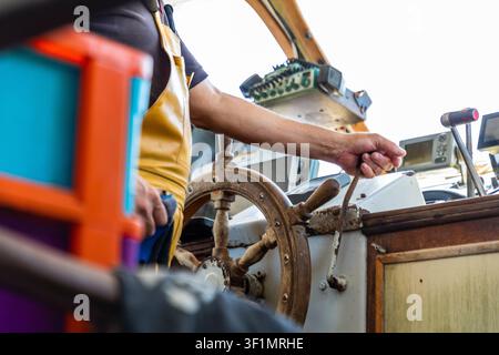 Nahaufnahme eines Fischers in Schürze, der ein kleines Fischerboot bedient, Hand am Gashebel und verwittertes Holzrad, mit Navigationsinstrumenten in der Kabine Stockfoto