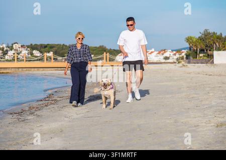 Ein fröhliches Paar genießt einen gemütlichen Spaziergang entlang eines sandigen Strandes, begleitet von ihrem verspielten Hund. Die sonnenbeschienene Küste bietet eine ruhige und malerische Stockfoto