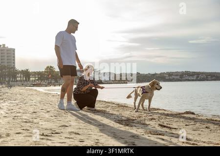Ein Paar genießt einen gemütlichen Spaziergang entlang des sandigen Strandes am Meer mit seinem Hund. Die ruhige Umgebung, sanfte Wellen und warme Sonne schaffen eine perfekte Stockfoto
