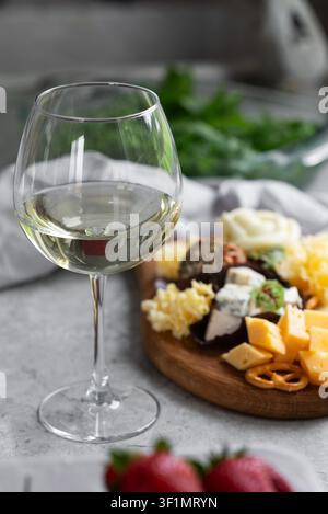 Elegante Nahaufnahme eines Glases Weißwein neben einem rustikalen Käsebrett mit verschiedenen Käsesorten, Nüssen und Obst, platziert auf einem hellen Tisch mit sanftem natürlichem L Stockfoto
