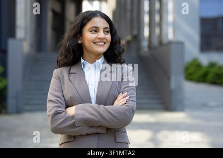 Selbstbewusste Geschäftsfrau lächelt mit verschränkten Armen, steht im Freien in einer modernen städtischen Umgebung und symbolisiert Karrierenerfolg, Ambition und berufliche Führung Stockfoto