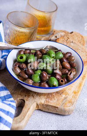Nahaufnahme von grünen und schwarzen Oliven in einer weißen Schüssel mit Olivenöl, serviert auf einem Holzbrett mit rustikalem Brot und Weingläsern, ein mediterranes Vergnügen Stockfoto