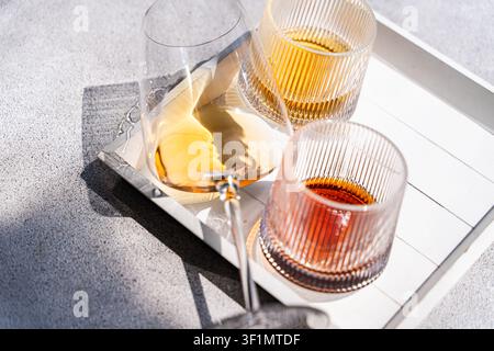 Oberblick auf gerippte Gläser mit bernsteinfarbenen Getränken auf einem weißen Tablett im hellen Sonnenlicht, eines ist umgekippt und verschüttet sich auf einer grauen Oberfläche, wodurch ein scharfer Schatten entsteht Stockfoto