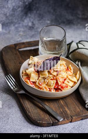 Rustikale Schüssel mit cremigen Nudeln in Tomatensauce, mit Basilikum und Pfeffer garniert, serviert auf einem Holzbrett mit Gabel, Leinenserviette und Wasserglas. Stockfoto