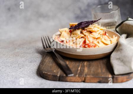 Nahaufnahme von cremigen Ravioli mit Tomatensauce und Basilikum in einer Keramikschüssel auf einem rustikalen Holzbrett, gestylt mit Gabel, Glas und Leinen auf grauem Untergrund Stockfoto