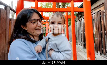 Ein freudiger Moment fängt eine Mutter ein, die warm lächelnd ihr Kind zwischen den lebhaften roten Torii-Toren des Horikoshi-Schreins in Osaka, Japan, betrachtet. Eine wunderschöne Mischung Stockfoto