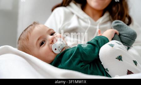 Ein fröhliches Baby genießt Qualitätszeit mit der Familie, gekleidet in festlicher Hose, die mit Weihnachtsbäumen verziert ist, und symbolisiert Wärme und Freude in einer gemütlichen Heimatszene Stockfoto