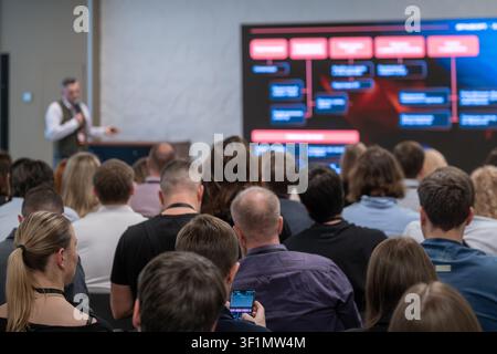 Konferenzpublikum hört dem Referenten zu, Reihen von Teilnehmern konzentrieren sich auf den großen Bildschirm, lockere Geschäftsumgebung, ein Teilnehmer verwendet ein Smartphone. Stockfoto