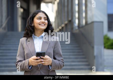 Junge professionelle Geschäftsfrau lächelt. Schaut nach vorne. Und hält ein Smartphone, während sie selbstbewusst im Freien vor einem modernen Firmengebäude steht Stockfoto