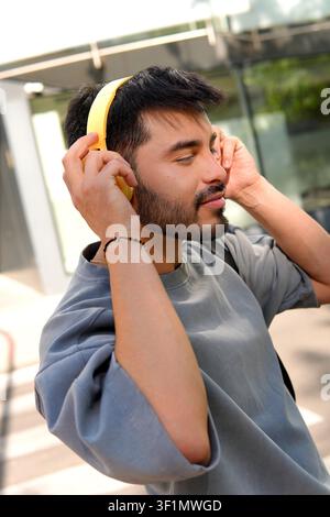 Junger Mann mit hellgelben Kopfhörern passt sie an, während er im Freien in einer städtischen Umgebung steht und während eines lockeren Spaziergangs Musik genießt, entspannt Stockfoto