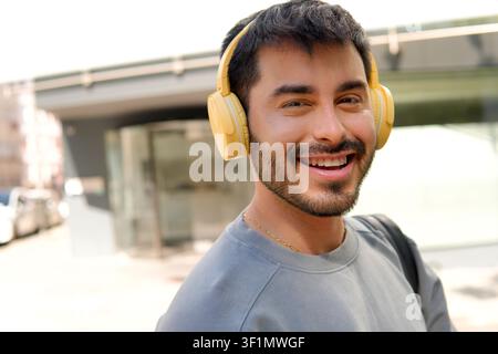 Nahaufnahmeporträt eines lächelnden jungen Mannes, der gelbe Kopfhörer trägt, im Freien auf einer sonnigen Straße der Stadt, einen Rucksack trägt und Musik genießt während eines... Stockfoto