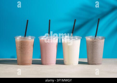 Vier Plastikbecher mit Eismilchshakes in verschiedenen Geschmacksrichtungen mit schwarzen Strohhalmen stehen in einer Reihe auf einem hellen Tisch vor einem hellblauen Hintergrund mit Stockfoto