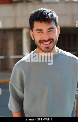 Nahaufnahme eines lächelnden jungen bärtigen Mannes, der einen grauen Sweatshirt trägt, steht im Freien im Sonnenlicht mit unscharfem urbanem Hintergrund, freundlicher Ausdruck Stockfoto