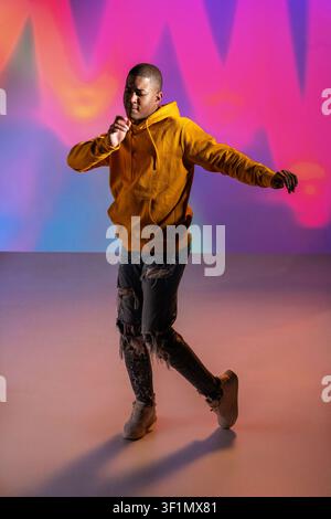 Schwarzer Mann, urbaner Tänzer in einem gelben Hoodie, führt eine dynamische Hip-Hop-Bewegung in einem Studio mit farbenfroher Neonbeleuchtung aus, moderner Streetwear-Stil und energisch Stockfoto