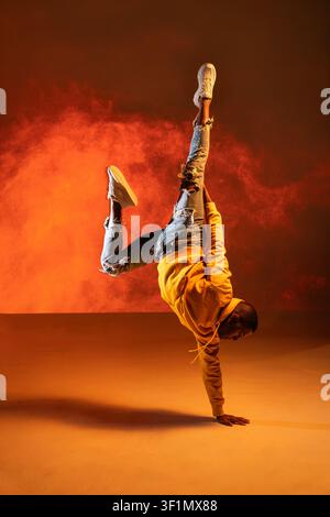 Schwarzer urbaner Tänzer führt im Studio einen kraftvollen einhändigen Handstand-Freeze aus, trägt Hoodie und Jeans, mit orangefarbenem Rauch und dramatischer Beleuchtung Stockfoto