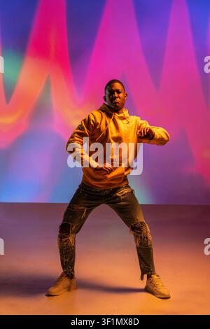 Schwarzer Mann, urbane Tänzer in einem gelben Hoodie, führt eine Hip-Hop-Bewegung in einem Studio aus, steht in dynamischer Pose vor einem lebendigen Neon-Farbverlaufshintergrund mit Stockfoto