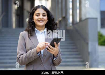 Junge professionelle Geschäftsfrau lächelt, schaut in die Kamera und interagiert mit einem Smartphone, während sie selbstbewusst vor einem modernen städtischen Gebäude steht Stockfoto