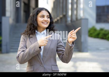 Junge Geschäftsfrau lächelt selbstbewusst, während sie im Freien vor einem Bürogebäude steht. Zeigt mit beiden Zeigefingern. Deutet auf Optionen. Werbung. Zukunftschancen Stockfoto