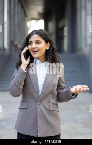 Junge indische Geschäftsfrau führt ein ausdrucksstarkes Telefongespräch. Lächelnd und gestikulierend während der Kommunikation im Freien in Berufskleidung. Repräsentiert Konzepte von Erfolg. Networking Stockfoto