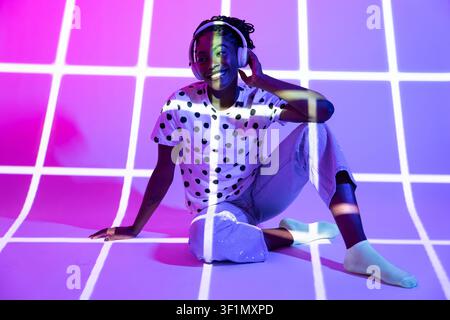 Lächelnde Afro-Frau Tänzerin mit Kopfhörern sitzt auf dem Studioboden in einem gepunkteten Hemd unter einer lila Neon-Gitter-Projektion, kreative Nachtleben-Stimmung, Musik Stockfoto