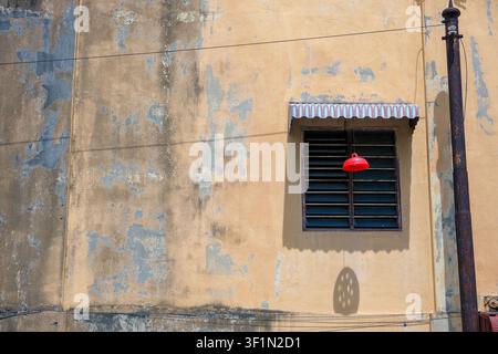 Eine verwitterte Denkmalwand mit einem Vintage-Fenster mit Lamellen, abblätternder Farbe und einer auffälligen roten Industrielampe in George Town, Penang, Malaysia. Stockfoto