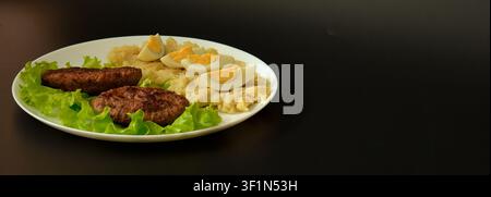 Zwei gebratene Fleischkoteletts auf einem Salatblatt, Kartoffelpüree und ein gekochtes Hühnerei auf einem weißen runden Keramikteller vor schwarzem Hintergrund. Nahaufnahme. Stockfoto