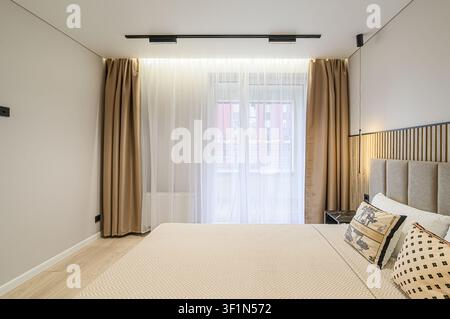 helle Schlafzimmerszene mit einem ordentlich gemachten Bett, hellbeigen Wänden, Holzfußboden und einem Fenster mit durchsichtigen und goldenen Vorhängen Stockfoto