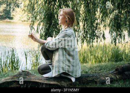 Frau in meditativer Haltung auf einem Baumstamm am See, Naturtherapiesitzung, Erdungstechniken, achtsames Atmen, Wohlbefinden im Freien Stockfoto