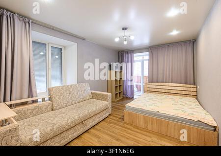 Gemütliches Zimmer mit einem gemusterten Sofa, einem Holzbett und helllila Vorhängen. Einfache Einrichtung, helle Beleuchtung. Stockfoto